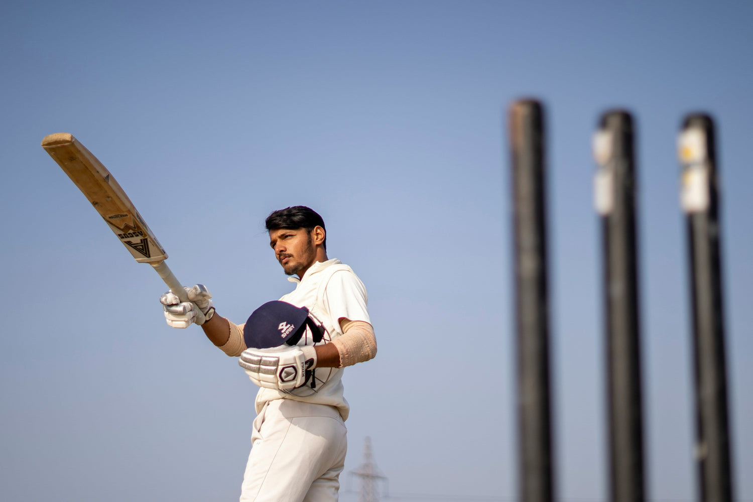 A young adult, Indian, posing with his cricket bat and helmet wearing his cricket uniform at a cricket field.